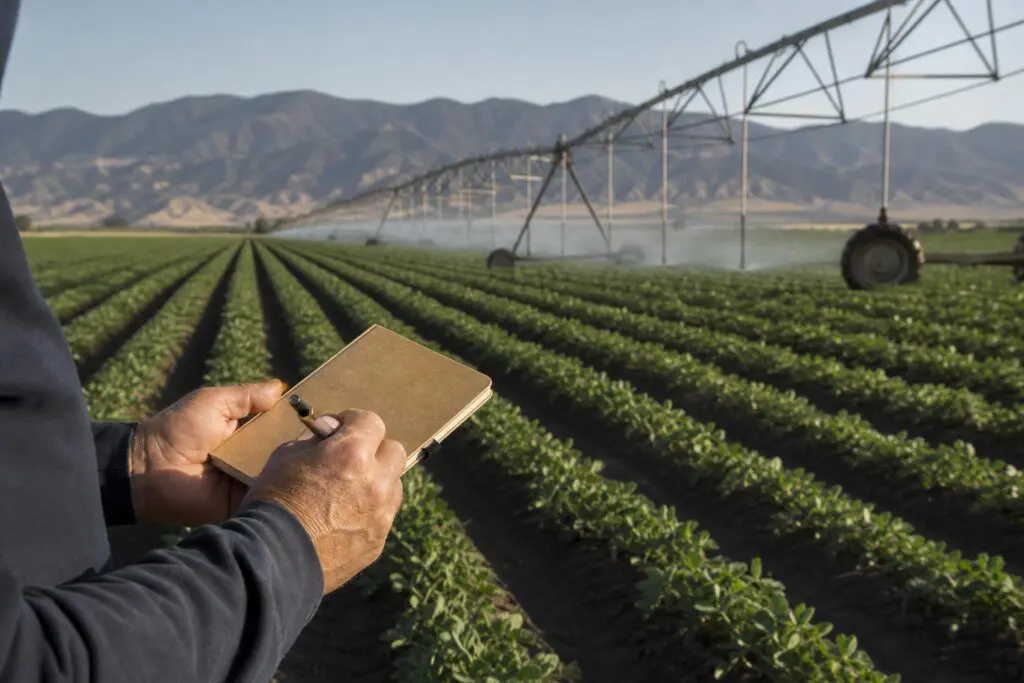 Hands holding a notebook beside crop rows and irrigation equipment.